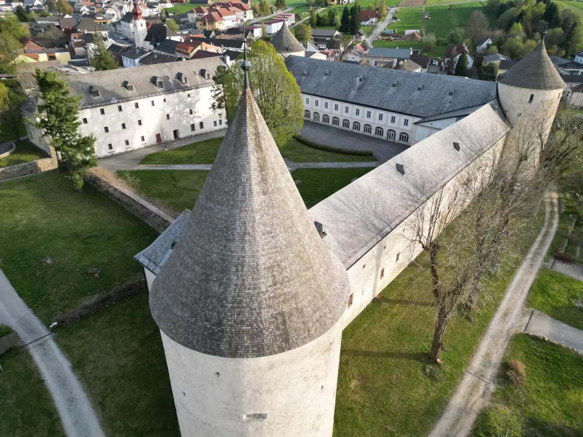 Eine Dronenaufnahme von einem Schloss, in der Mitte vom Bild ist ein Turm mit kegelförmigen Dach. Der Innenhof ist schön zu sehen und im Hintergrund befindet sich die Ortschaft Ottenschlag